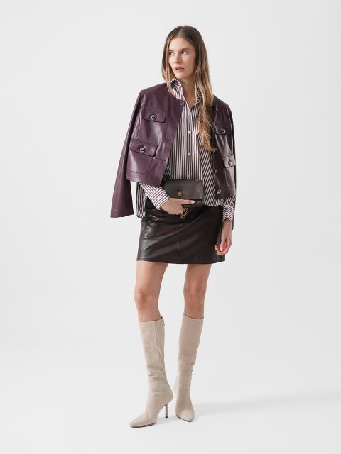 Eine Frau posiert in einer kastanienbraunen Lederjacke, einem gestreiften Hemd, einem kurzen Lederrock, beigefarbenen kniehohen Stiefeln und hält eine schwarze Clutch vor einem weißen Hintergrund.