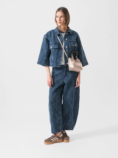 Eine Frau, die eine blaue Jeansjacke und weite Jeans, braune Turnschuhe und eine Metallhandtasche trägt, steht vor einem schlichten weißen Hintergrund.