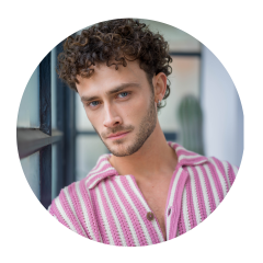 Mann mit lockigen braunen Haaren und blauen Augen, der ein rosa-weiß gestreiftes Hemd trägt und in einem Innenraum an einem Fenster in die Kamera schaut.