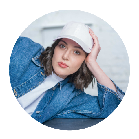 Frau mit weißer Mütze und Jeansjacke, die sich auf ihren Arm stützt und mit neutralem Blick in die Kamera schaut.