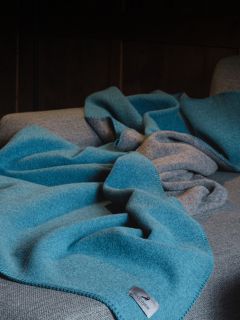 Eine blaue Wolldecke mit grauer Unterseite ist über einem grauen Sofa drapiert, wobei im Vordergrund ein Stoffetikett zu sehen ist.