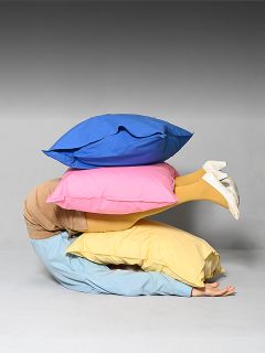 Eine Person liegt zusammengerollt auf dem Boden, zugedeckt von drei großen bunten Kissen, aus denen nur ihre gelb gekleideten Beine und weißen Schuhe herausragen.