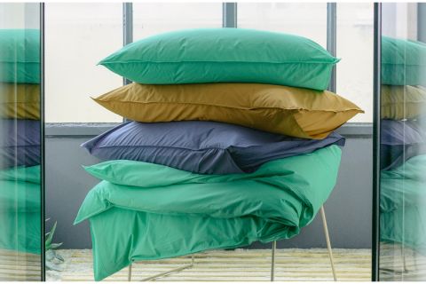 Vier farbenfrohe Kissen, grün, senfgelb, marineblau und blaugrün, sind auf einem Stuhl vor großen Fenstern im Industriestil gestapelt.