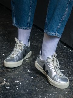 Person in silbernen Metallic-Turnschuhen mit weißen Sohlen, weißen Socken und blauen Jeans, die auf einem dunklen, strukturierten Boden steht.