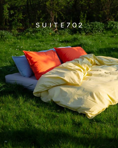 Ein Bett mit leuchtend orangefarbenen und roten Kissen und einer gelben Bettdecke steht im Freien auf grünem Gras im Sonnenlicht. Darüber steht "SUITE702".