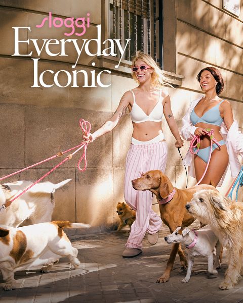 Zwei lächelnde Frauen in Unterwäsche gehen mit mehreren Hunden auf einer sonnigen Straße in der Stadt spazieren. Der Text lautet: "Sloggi Everyday Iconic.