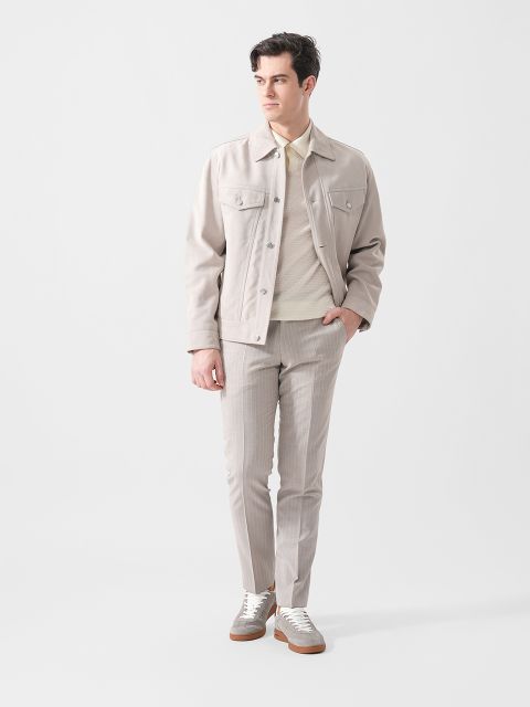 Ein Mann, der eine hellbeige Jacke, einen cremefarbenen Pullover, eine hellgraue Hose und weiße Turnschuhe trägt, steht vor einem schlichten weißen Hintergrund.