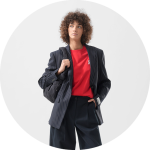 Frau mit lockigem Haar trägt einen Nadelstreifenblazer, ein rotes Hemd und eine schwarze Umhängetasche und posiert mit einer Hand an der Hüfte vor einem hellen Hintergrund.