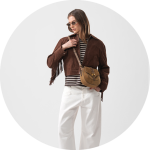 Frau mit Sonnenbrille, brauner Fransenjacke, gestreiftem Hemd, weißer Hose und einer kleinen hellbraunen Umhängetasche in der Hand vor einem hellen Hintergrund.