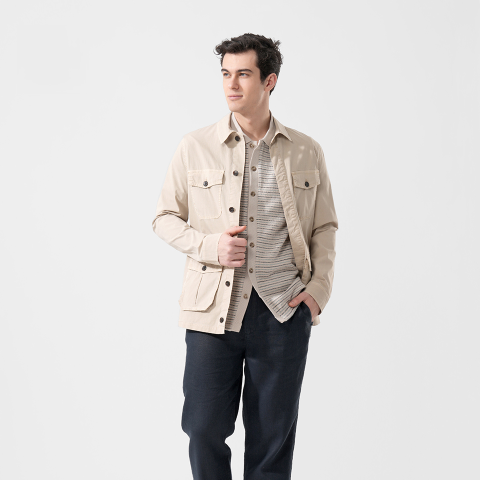 Ein Mann in einer beigen Jacke über einem gestreiften Hemd und einer dunklen Hose steht mit einer Hand in der Tasche und blickt vor einem weißen Hintergrund zur Seite.