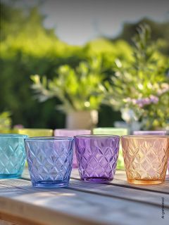 Vier strukturierte, bunte Glasbecher stehen auf einem Holztisch im Freien, mit unscharfen grünen Pflanzen und Sonnenlicht im Hintergrund.