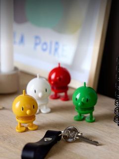 Vier bunte, runde Spielzeugfiguren stehen auf einem Schreibtisch neben Schlüsseln und einem Notizbuch, mit einem unscharfen gerahmten Kunstwerk im Hintergrund.