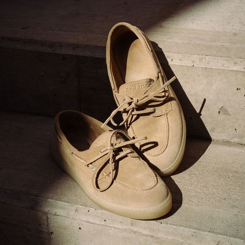 Ein Paar beigefarbene Bootsschuhe aus Wildleder mit Schnürsenkeln, die auf Betonstufen in teilweisem Sonnenlicht stehen.