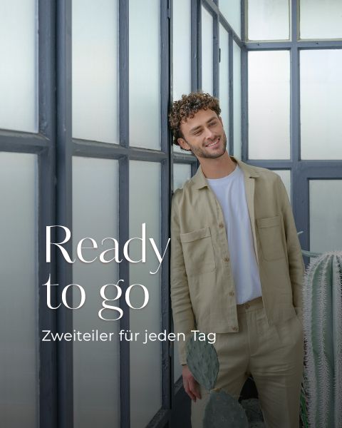 Ein Mann in hellbeiger Kleidung steht vor großen Milchglasfenstern und lächelt. Der Text lautet: "Bereit zu gehen. Zweiteiler für jeden Tag.
