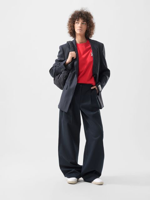 Person mit lockigem Haar trägt einen Nadelstreifenblazer, ein rotes Hemd, eine Hose mit weitem Bein, weiße Schuhe und eine schwarze Tasche vor einem schlichten Hintergrund.