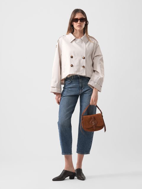 Eine Frau mit Sonnenbrille trägt eine beigefarbene doppelreihige Jacke, eine abgeschnittene blaue Jeans, schwarze Schuhe und hält eine braune Handtasche vor einem unifarbenen Hintergrund.