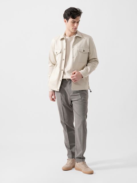 Mann in cremefarbener Jacke, weißem Hemd, grauer Hose und beigen Turnschuhen, vor einem schlichten weißen Hintergrund.