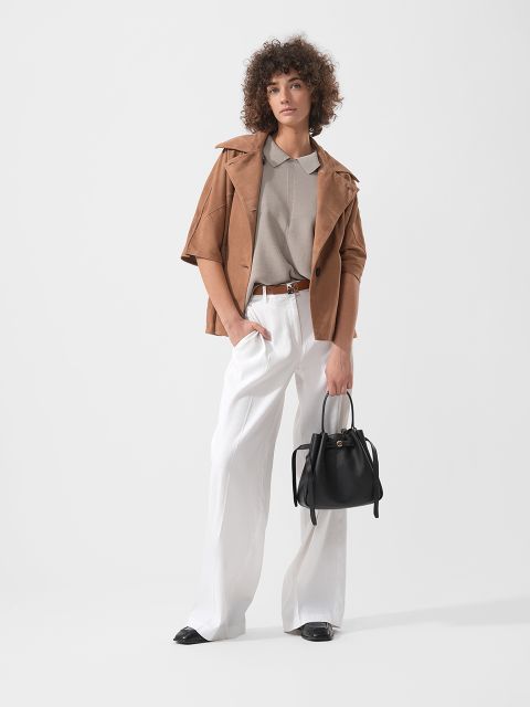 Frau mit lockigem Haar, die eine hellbraune Jacke, ein beiges Hemd und eine weiße Hose mit weitem Bein trägt und eine schwarze Handtasche in der Hand hält, steht vor einem schlichten Hintergrund.