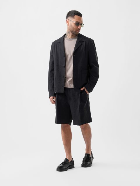 Ein Mann mit Sonnenbrille, schwarzer strukturierter Jacke, beigem T-Shirt, schwarzen Shorts und schwarzen Slippern steht vor einem schlichten weißen Hintergrund.