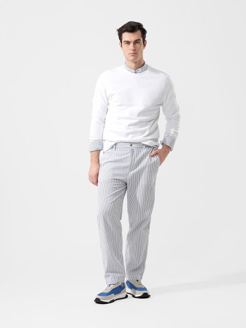 Ein Mann steht vor einem schlichten Hintergrund und trägt ein weißes Sweatshirt, eine hellblau gestreifte Hose und blau-weiße Turnschuhe.