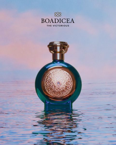 Ein blau-goldener „Boadicea“-Parfümflakon steht auf spiegelglattem Wasser vor dem Hintergrund eines pastellfarbenen Himmels.