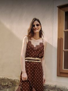 Eine Frau in einem braunen gepunkteten Kleid mit Spitzenbesatz, Sonnenbrille und Goldschmuck steht lächelnd vor einer beigen Wand und einem Fenster.