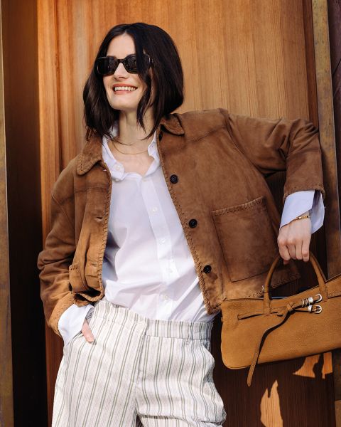 Frau mit Sonnenbrille, die eine braune Jacke, ein weißes Hemd und eine gestreifte Hose trägt, lächelt und eine braune Handtasche vor einem hölzernen Hintergrund hält.
