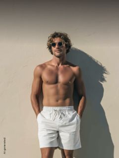 Ein Mann ohne Hemd, mit Sonnenbrille und gestreiften Shorts steht vor einer sonnenbeschienenen Wand, die Hände in den Taschen, und wirft einen Schatten.