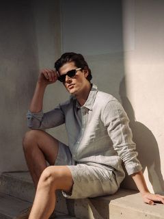 Ein Mann mit Sonnenbrille, hellgrauem Button-up-Hemd und passenden Shorts sitzt auf einer Betontreppe im Sonnenlicht vor einer hellen Wand.