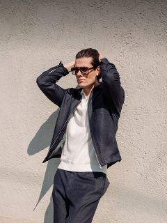 Ein Mann mit Sonnenbrille, dunkler Jacke und dunkler Hose steht an einer strukturierten Wand, die Hände hinter dem Kopf, und schaut in die Ferne.