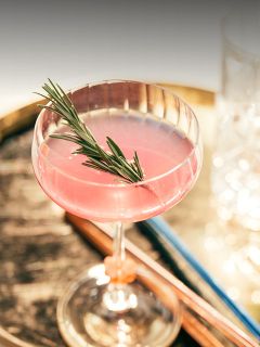 Ein rosafarbener Cocktail, garniert mit einem Rosmarinzweig in einem Coupe-Glas, auf einer rustikalen Holzfläche mit verschwommenen Gläsern im Hintergrund.