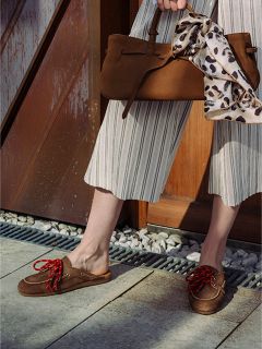 Person, die eine gestreifte Hose mit weitem Bein und braune Slipper mit roten Schnürsenkeln trägt und eine braune Handtasche in der Hand hält, an deren Griff ein Schal mit Leopardenmuster gebunden ist.