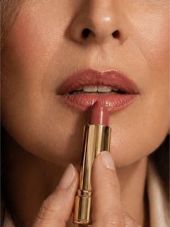 Nahaufnahme einer Frau, die mit einer goldenen Lippenstifttube lila Lippenstift auf ihre Lippen aufträgt.