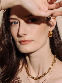 Eine Frau mit heller Haut und braunem Haar, die Goldschmuck trägt, posiert mit ihrer Hand, die ihr Gesicht teilweise vor dem Licht schützt.