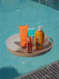 Ein Tablett mit vier Sonnenschutzmitteln steht neben einem Schwimmbad, wobei das Wasser und der Beckenrand im Hintergrund zu sehen sind.