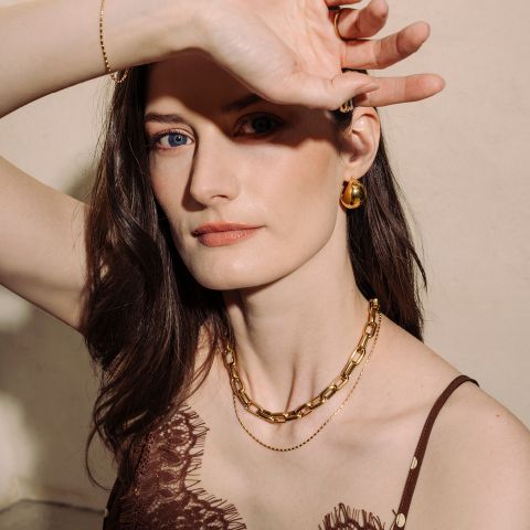 Frau mit heller Haut, die Goldschmuck und ein braunes Spitzenoberteil trägt und mit einem erhobenen Arm posiert, um ihr Gesicht teilweise vor dem Licht zu schützen.