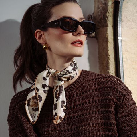 Frau mit schwarzer Sonnenbrille, goldenen Ohrringen, einem braunen Strickoberteil und einem gemusterten Schal, der um den Hals gebunden ist, steht im Sonnenlicht.