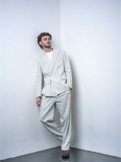 Ein Mann mit lockigem Haar, der einen hellgrauen Anzug und ein weißes T-Shirt trägt, lehnt an einer weißen Wand in einer minimalistischen Studioumgebung.