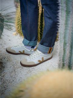 Person in Jeans mit Stulpen und weißen Turnschuhen mit beigen Akzenten, steht auf sandigem Boden zwischen hohen grünen Kakteen.