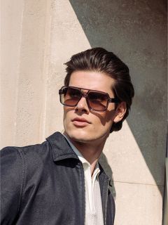 Mann mit gestyltem braunem Haar trägt eine große quadratische Sonnenbrille, eine dunkle Jacke und ein helles Hemd und steht im Sonnenlicht neben einer strukturierten Wand.