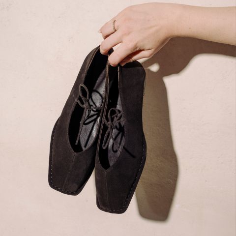 Eine Hand hält ein Paar schwarze Wildlederschnürschuhe mit quadratischen Zehen vor einer hellbeigen, strukturierten Wand.