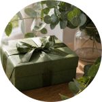 Auf einem Holztisch steht eine grüne Geschenkschachtel mit einer passenden Schleife neben einer Vase mit grünen Zweigen.