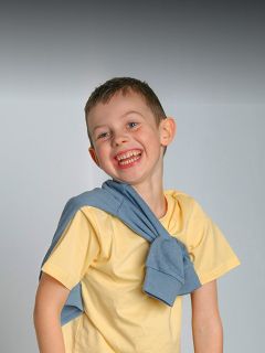 Lächelnder Junge in einem gelben Hemd mit einem blauen Pullover über den Schultern, der vor einem schlichten grauen Hintergrund steht.