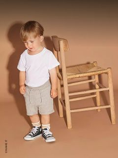 Ein junges Kind in einem weißen T-Shirt, grauen Shorts und schwarzen Turnschuhen steht neben einem Holzstuhl vor einem beigen Hintergrund.
