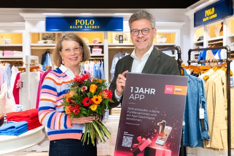 Zwei lächelnde Menschen stehen in einem Bekleidungsgeschäft; einer hält Blumen in der Hand, der andere ein Schild über eine einjährige App-Aktion.