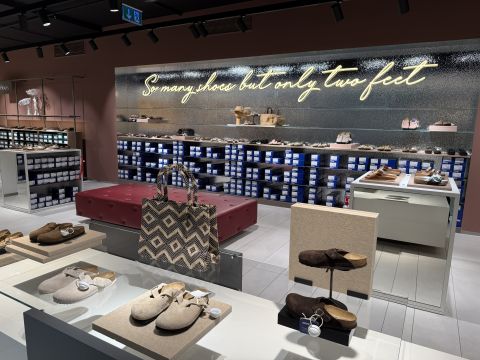 Interieur eines Schuhgeschäfts mit ausgestellten Sandalen, einer gemusterten Tragetasche und einer Leuchtreklame mit der Aufschrift "So viele Schuhe, aber nur zwei Füße".
