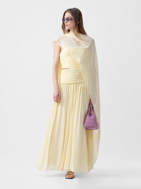 Frau in einem fließenden blassgelben Kleid mit drapierter Schulter, Sonnenbrille, Sandalen und einer kleinen lila Handtasche vor einem schlichten Hintergrund.