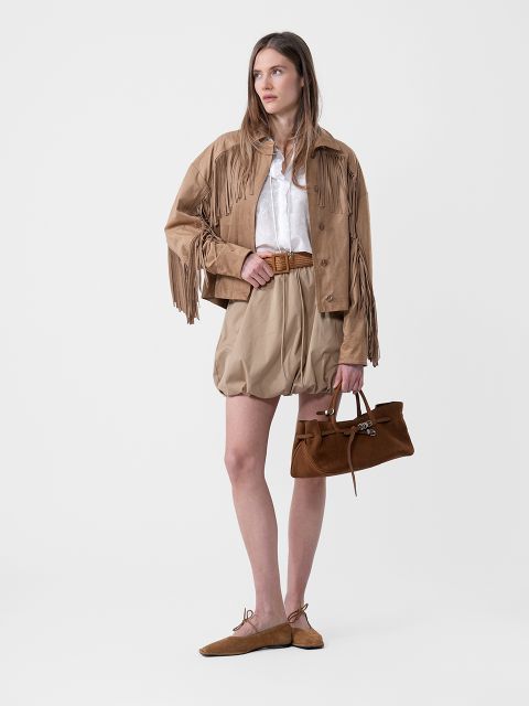 Frau in hellbrauner Fransenjacke, weißer Bluse, hellbraunen Shorts mit Gürtel, braunen Flats und einer braunen Handtasche vor einem schlichten Hintergrund.