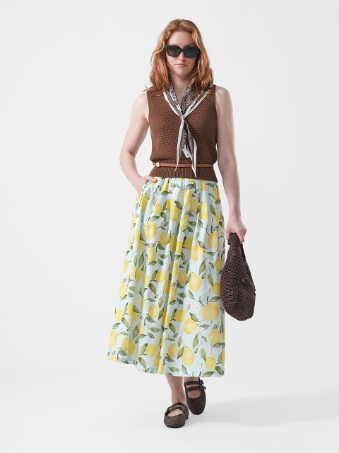 Frau mit Sonnenbrille, braunem ärmellosen Oberteil und langem Rock mit Zitronendruck, die eine braune Tasche in der Hand hält und einen Schal um den Hals trägt, steht vor einem weißen Hintergrund.