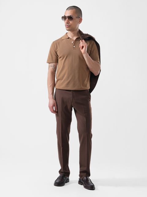 Ein Mann steht vor einem weißen Hintergrund, trägt eine Sonnenbrille, ein braunes Polohemd, eine dunkelbraune Hose und hält eine dunkle Jacke über der Schulter.